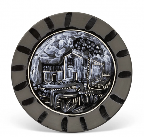 A ceramic plate by Pablo Picasso featuring a black and white painted rustic landscape with houses, foliage, and clouds, surrounded by a dark rim with vertical brushstroke patterns.