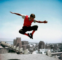 Frank Worth, Sammy Davis Jr in Mid-Air, photograph capturing Sammy Davis Jr. leaping on a rooftop with a cityscape background, exuding charisma.