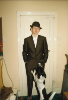 Nan Goldin Mod Kid With Dog photographic c-print showing boy in suit with hat and dog jumping up in a small domestic room.