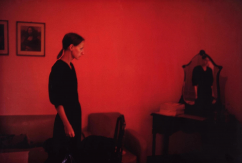 Nan Goldin Suzanne With Mona Lisa c-print showing woman in red-lit room with faint reflection in mirror and Mona Lisa on wall.